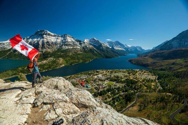 Das Foto zeigt eine beeindruckende Berg- und Seenlandschaft in Kanada. Ein Mann steht mit kanadischer Flagge auf einem Felsen und schaut auf eine Kleinstadt im Tal herab.