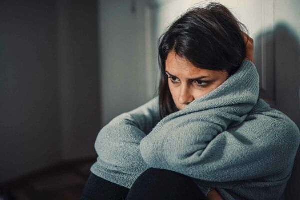 Das Foto zeigt eine sitzende Frau, die ihren Kopf in den Armen vergräbt und traurig und gedankenverloren ins Leere blickt.