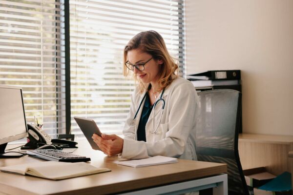 Eine junge blonde Ärztin mit Brille und Stethoskop um den Hals sitzt an einem Schreibtisch und schaut zufrieden auf das Tablet n ihrer Hand.