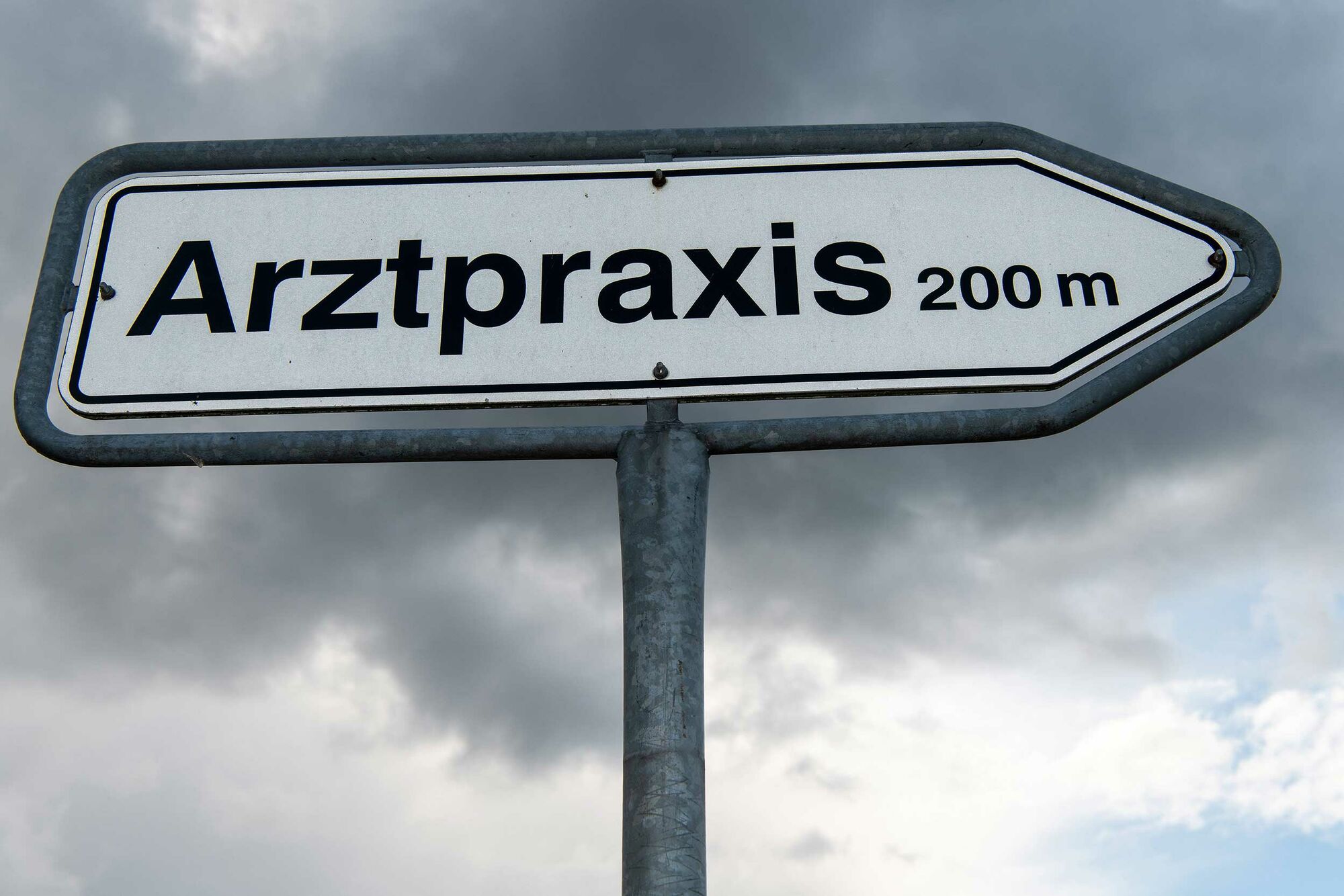 Das Foto zeigt ein Hinweisschild vor wolkenverhangenem Himmel. Auf dem Schild steht „Arztpraxis 200 Meter“.