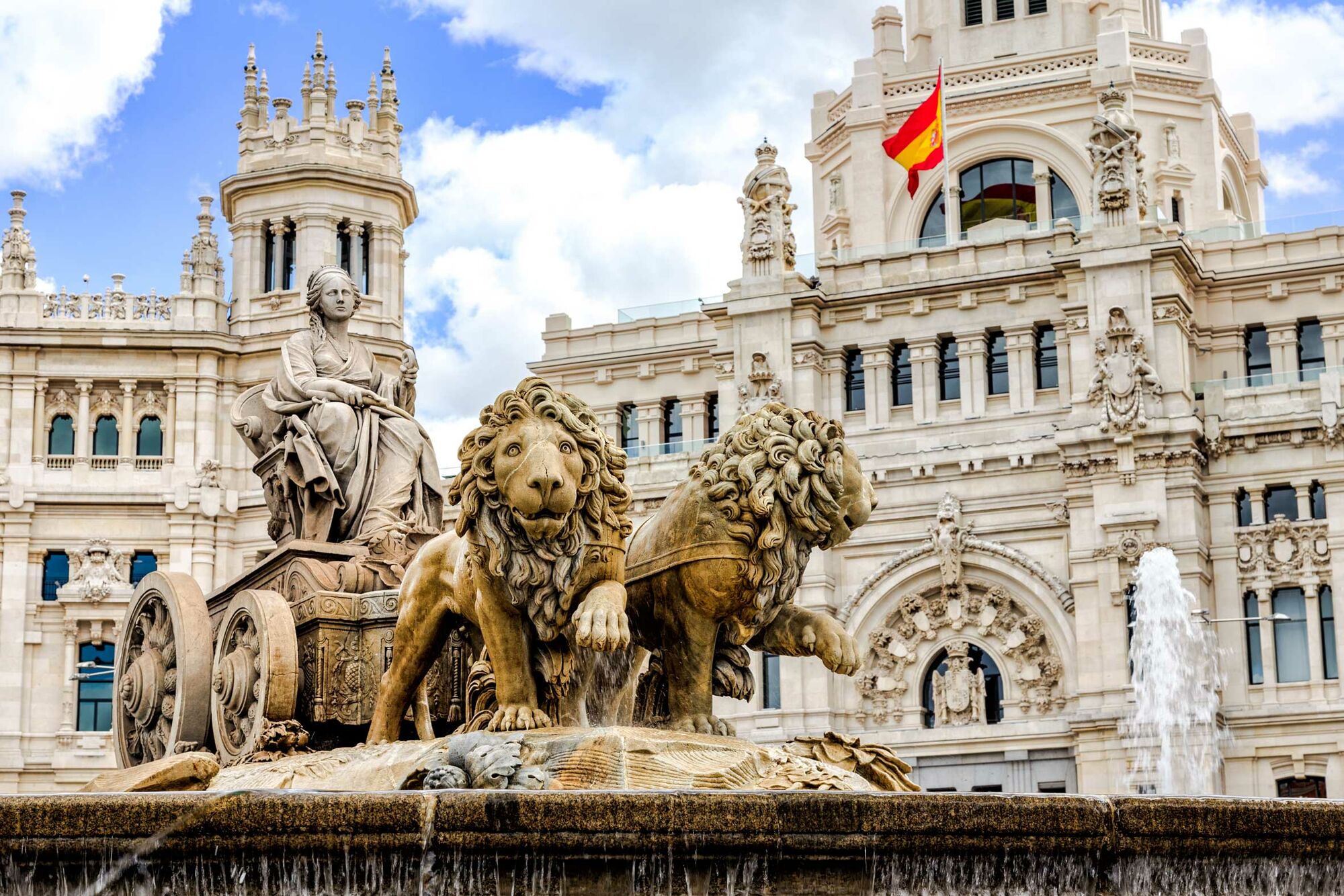 Das Bild zeigt den Spirngbrunnen Fuente de Cibeles in Madrid. Im Hintergrund ist der Palacio de Cibeles, der Sitz der Stadtverwaltung, zu sehen, an dem die Flagge Spaniens weht.