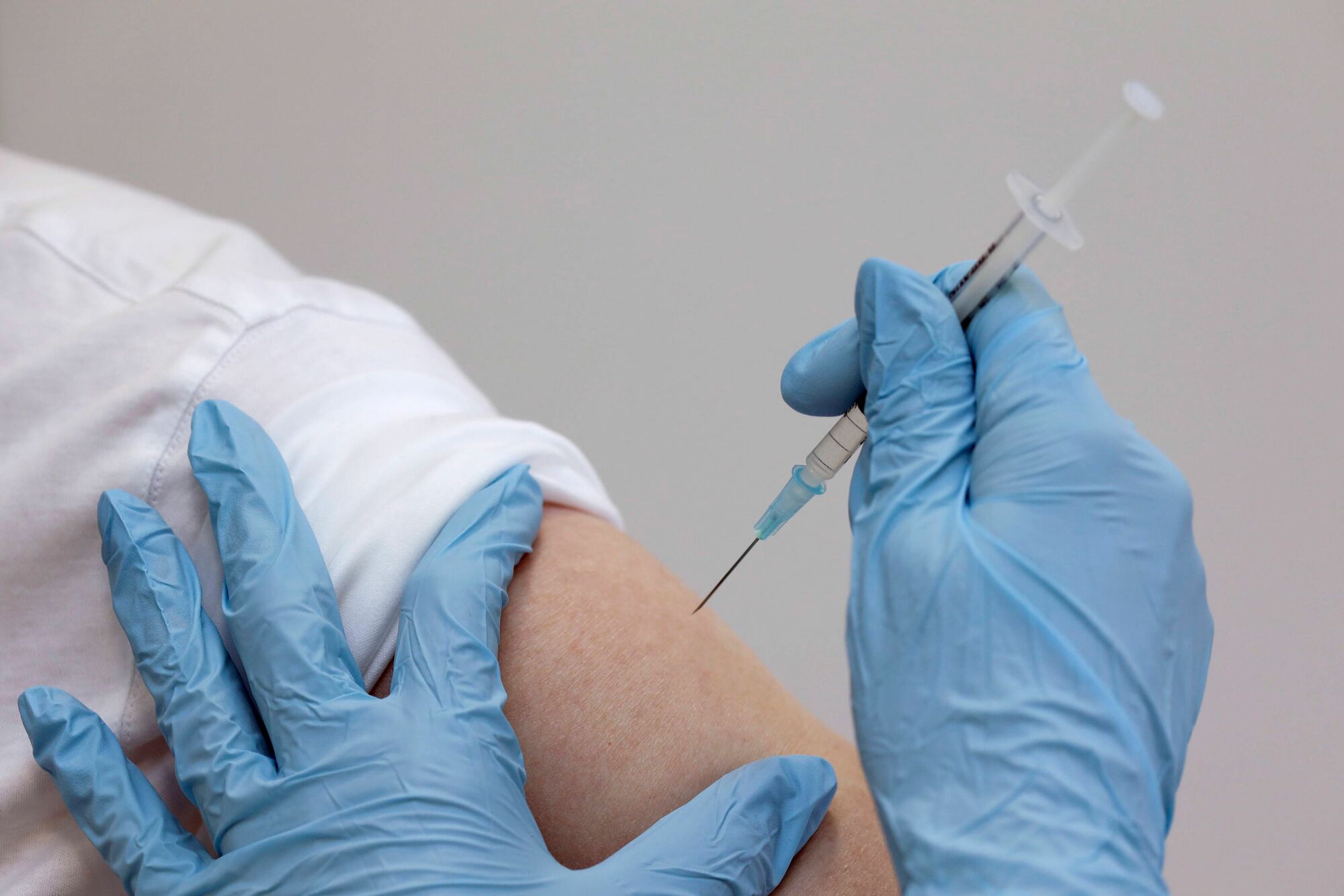 Das Foto zeigt den Oberarm eines Patienten, zwei Hände in blauen, medizinischen Einmalhandschuhen, die unmittelbar davor sind, die Kanüle einer Spritze in den Arm einzuführen.