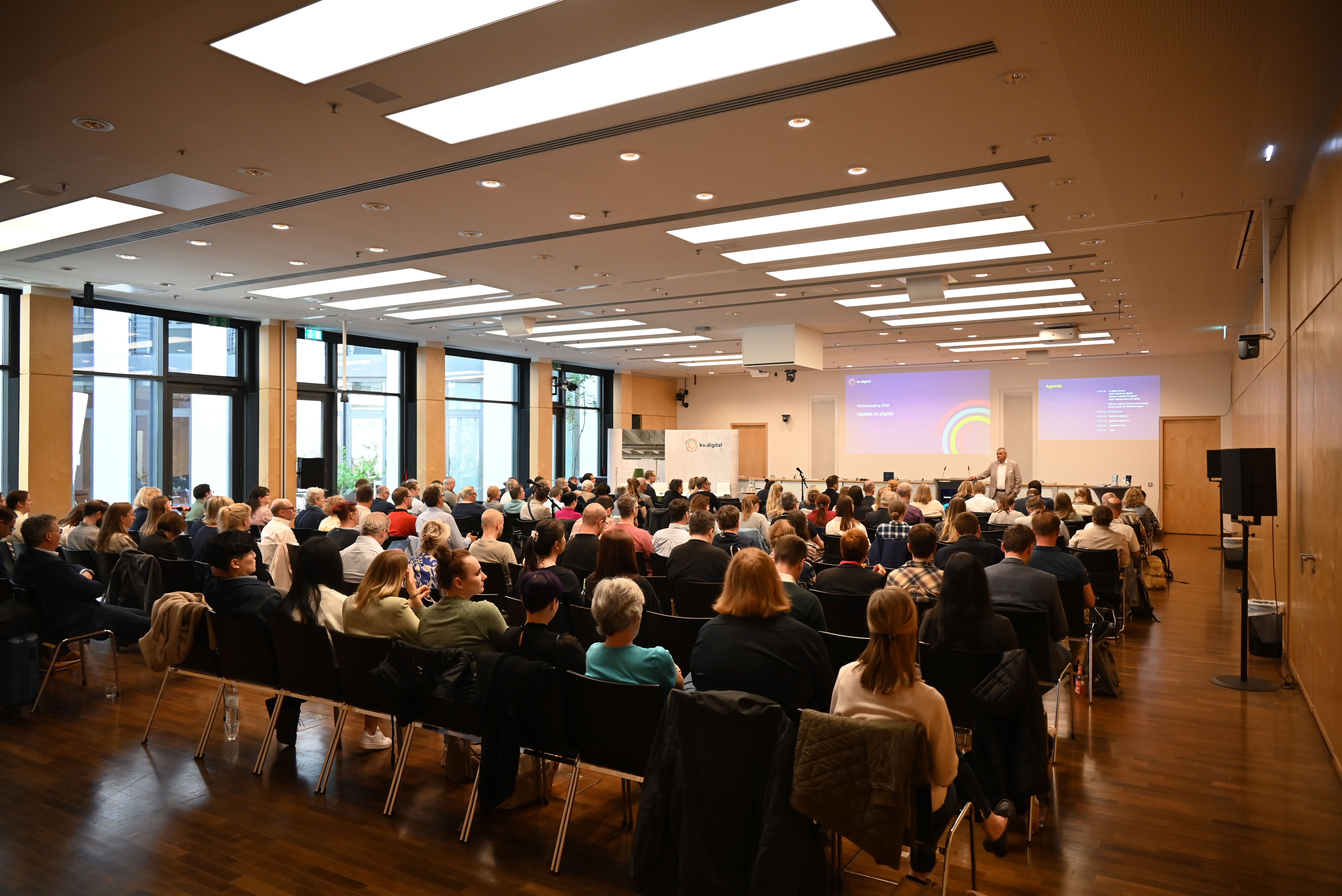 Ein großer Saal voller Menschen, die auf Stühlen in Reihen sitzen und gemeinsam einer Präsentation auf einer Leinwand folgen.